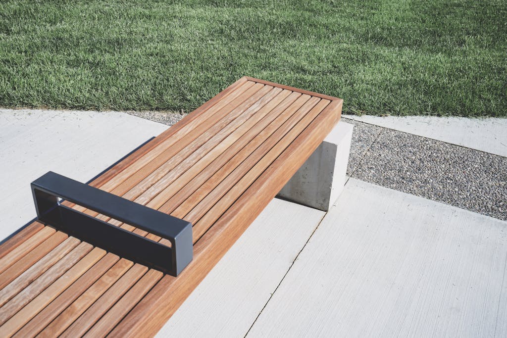 A minimalist wooden bench placed on a concrete path beside a green lawn in daylight.